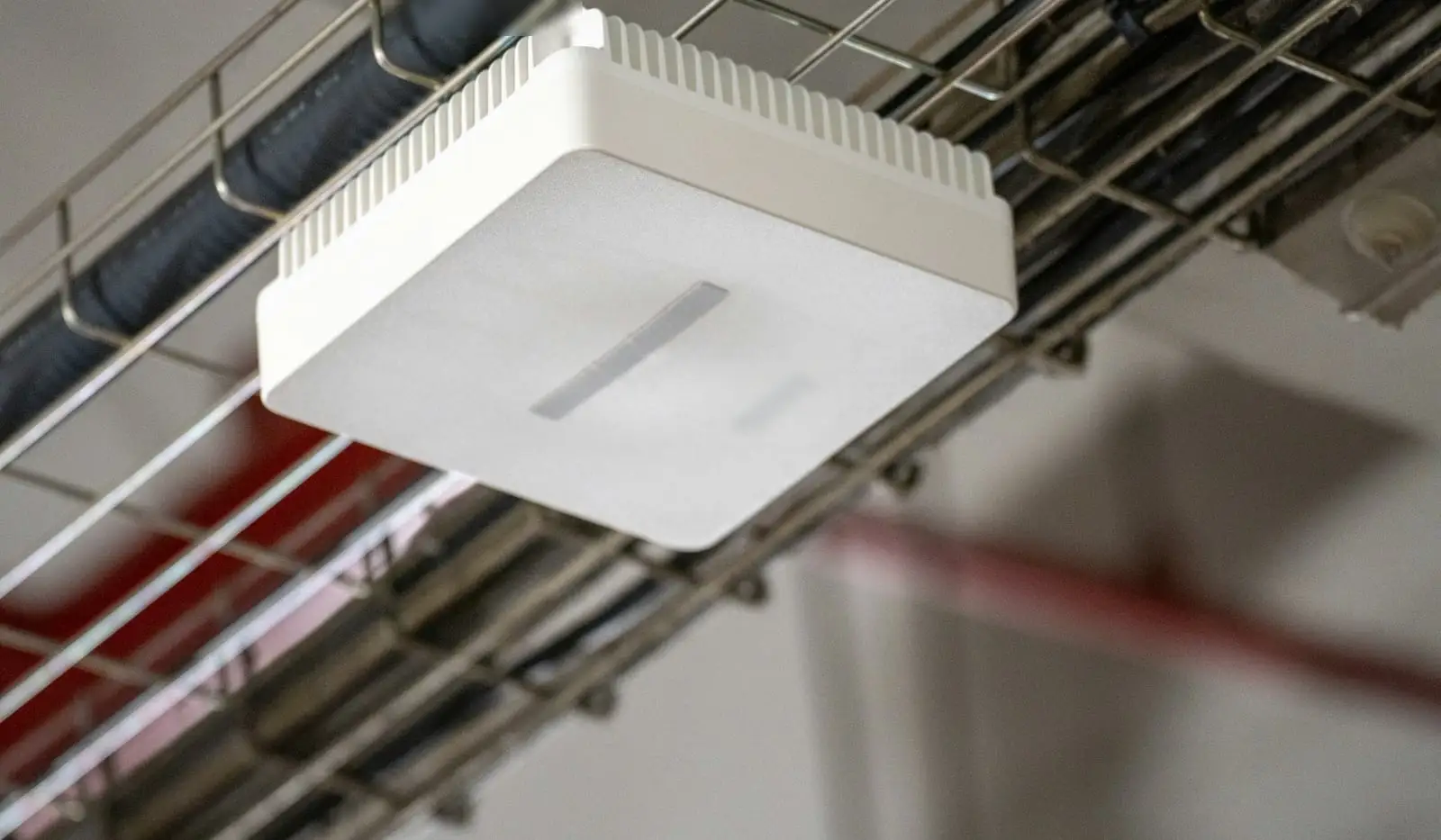 A white Wi-Fi access point hanging from a wire cable tray under a concrete ceiling.