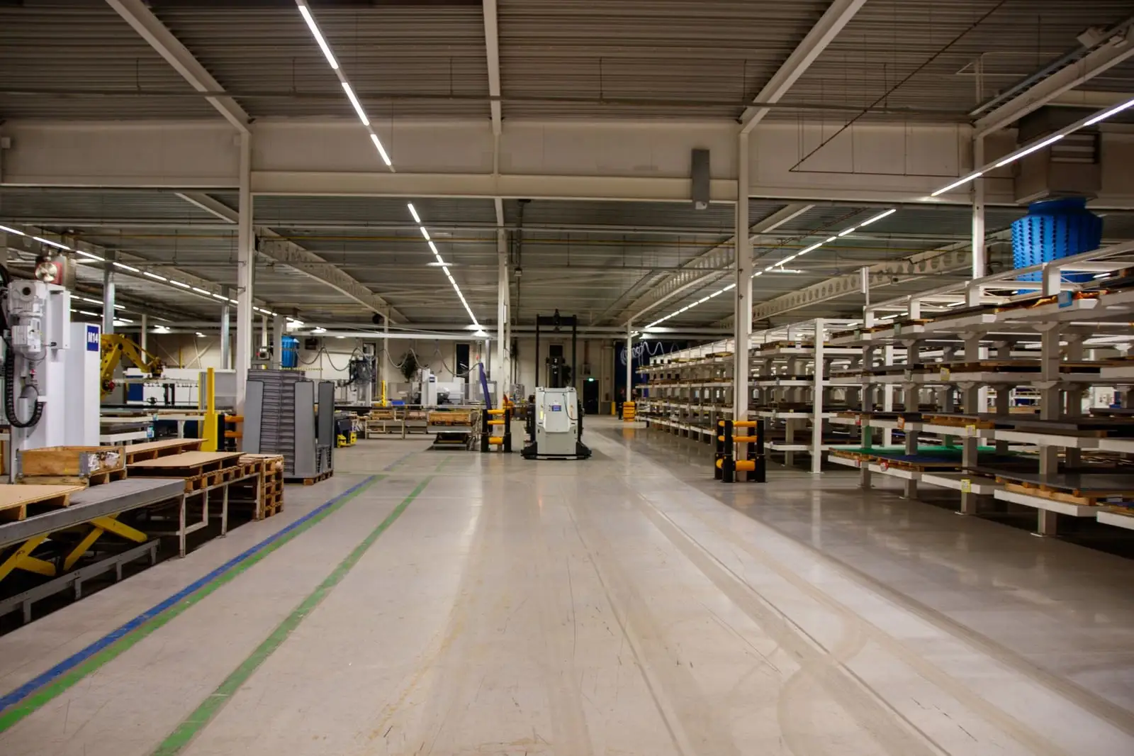 A wide factory hall under rows of lights, with long storage racks on the right and a pallet-handling vehicle in the aisle.