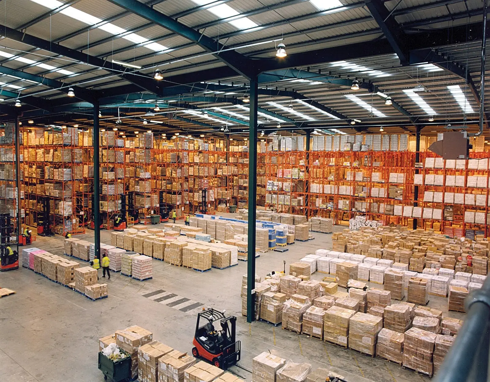 A large warehouse with long rows of steel pallet racking stacked with goods, and a forklift in the foreground.