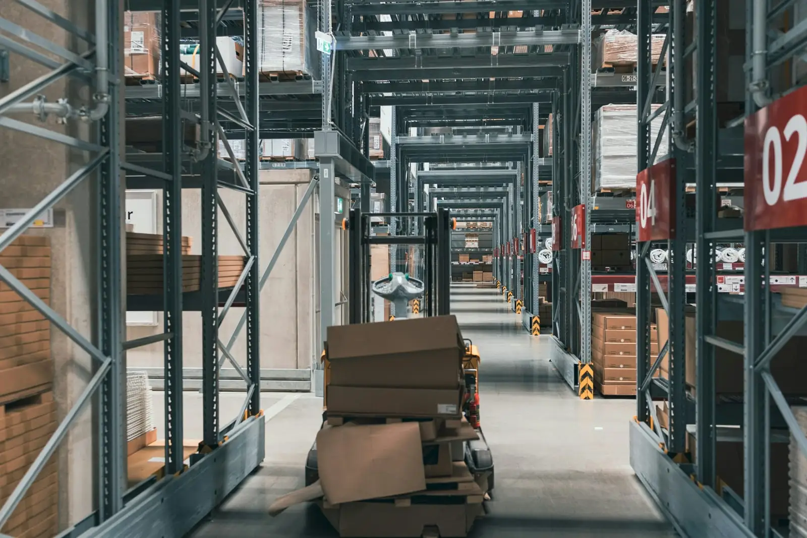 A pallet truck loaded with cardboard boxes in a narrow aisle between tall steel racks.