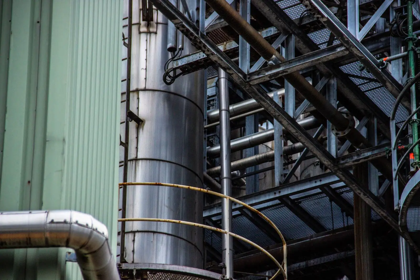 A tall metal vessel with a curved handrail at its base, beside a dense steel frame of pipes, grated walkways and cable tray, with green wall cladding on the left.