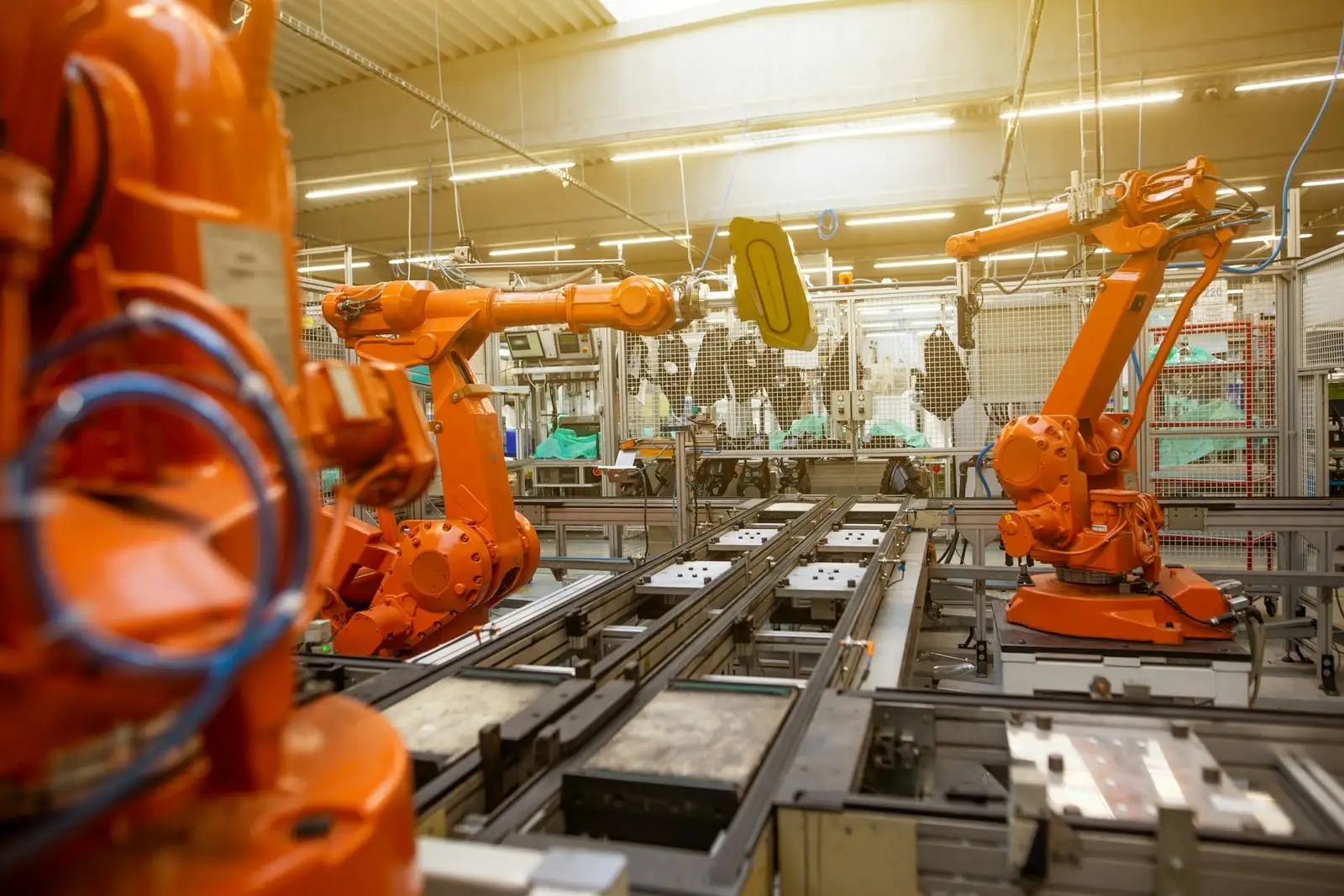 Orange industrial robot arms at a station on an assembly line, with conveyors and guarding around them.
